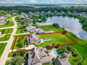 Aerial view of a Houston neighborhood highlighting a property with a red outline. Includes a large house, spacious yard, swimming pool, and nearby lake. Real estate showcasing in a scenic suburban setting.