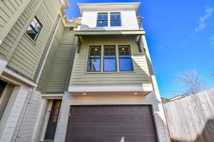 Modern multi-story townhouse in Houston with green siding, large windows, and a two-car garage viewed from below, showcasing contemporary architecture under a clear blue sky.