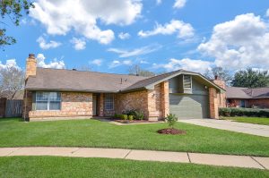 Single-story brick house with a gray garage in Houston, featuring a well-maintained lawn and clear blue skies. Ideal for real estate and video tours showcasing Houston realtor photography.