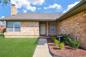 Front view of a single-story brick home with a well-maintained lawn and landscaped garden, highlighting the entry pathway leading to a wooden front door in Houston, ideal for real estate photography and video tour promotions.