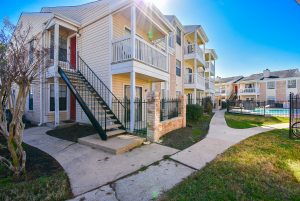 Apartment complex exterior with balconies and staircases, featuring a swimming pool area in a Houston real estate setting, clear blue sky.